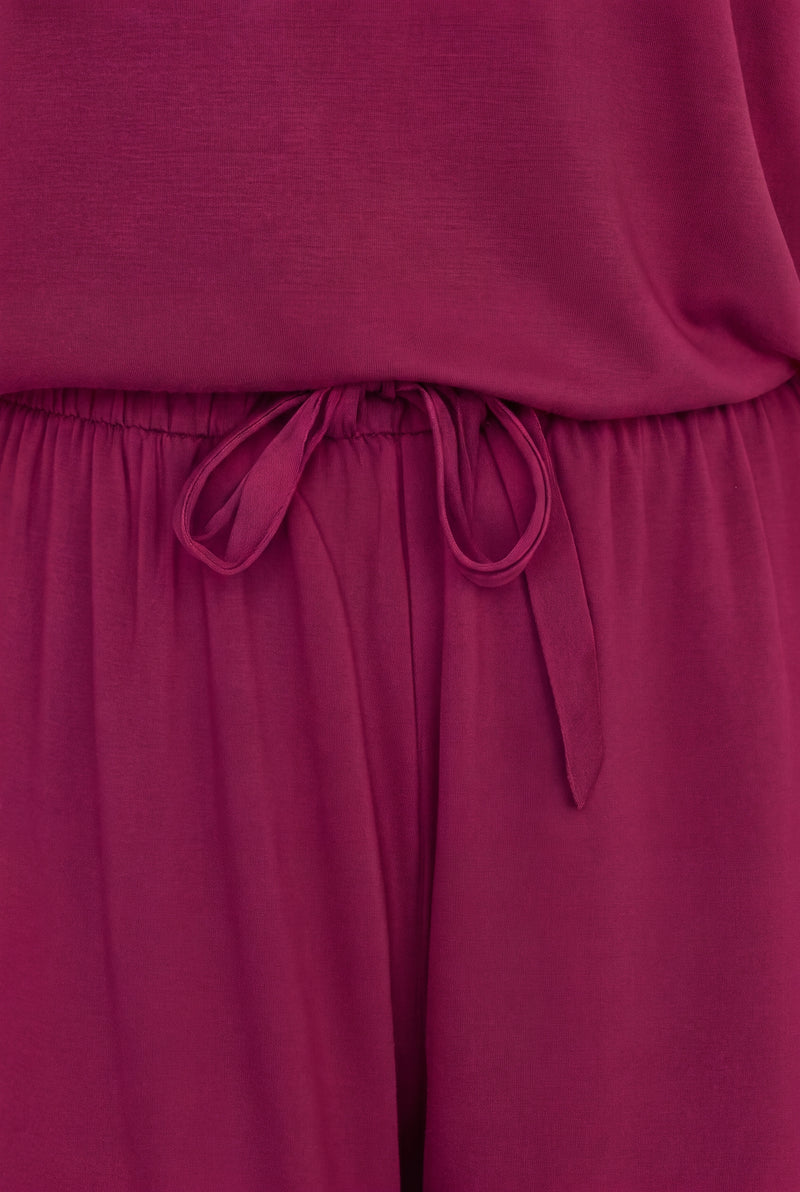 A close-up of a magenta-purple jumpsuit or romper, showing the waistband with a matching fabric drawstring tie and the flowing fabric of the pants below.