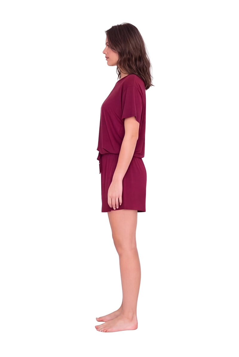 A woman stands in profile wearing a matching short-sleeve top and shorts set in a deep burgundy or maroon color, photographed against a plain light studio background.